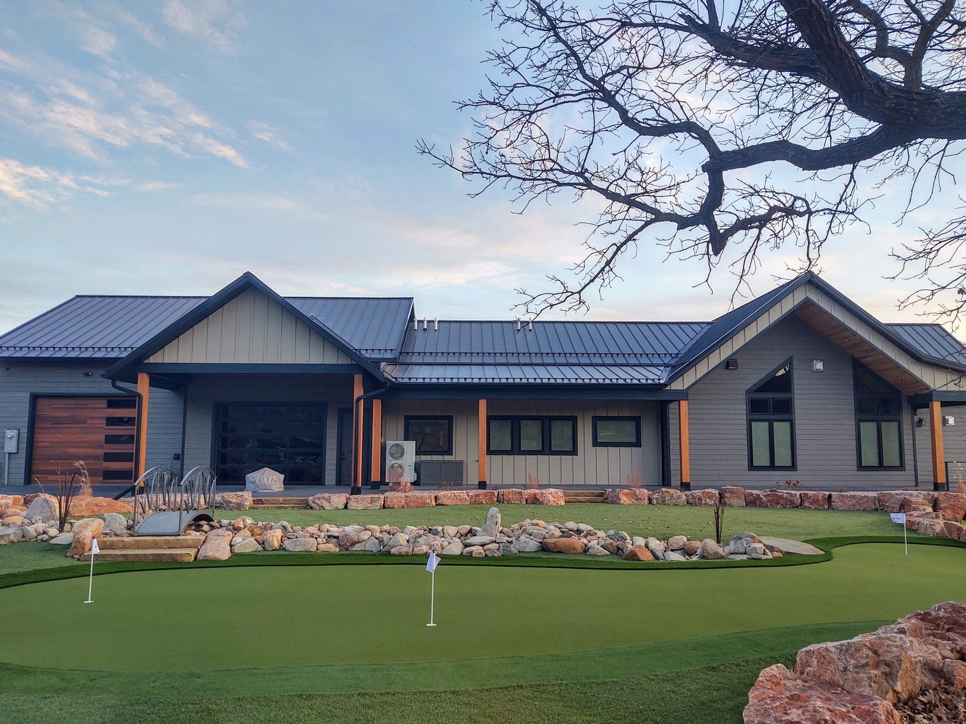 A gray, modern house with a metal roof and wooden accents, featuring an outdoor putting green in the foreground.