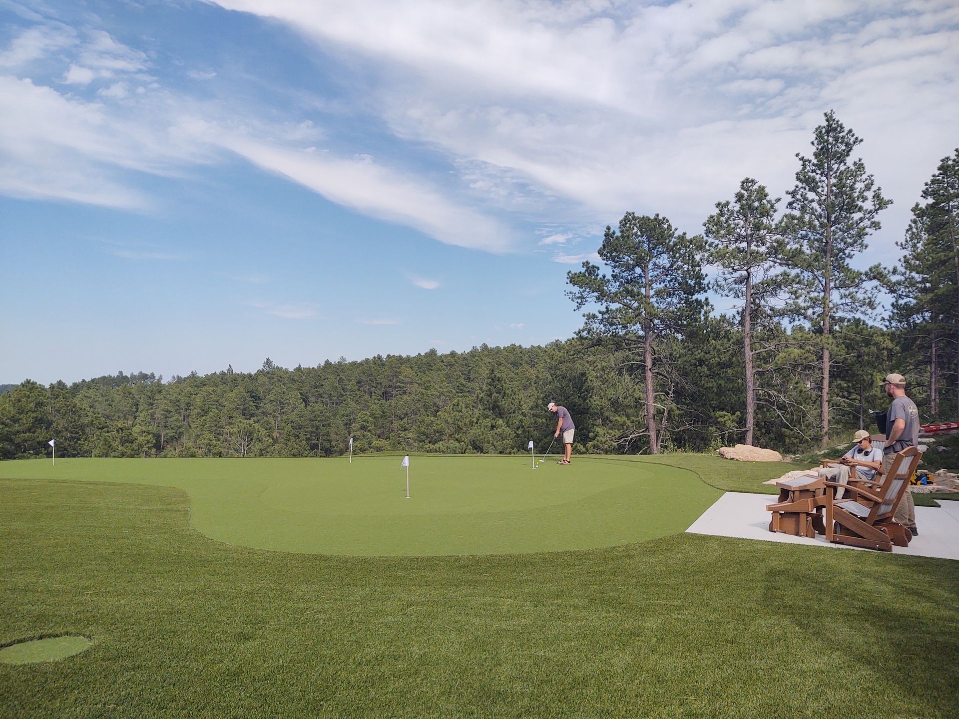 Man putting golf ball on a green, forested hillside, blue sky