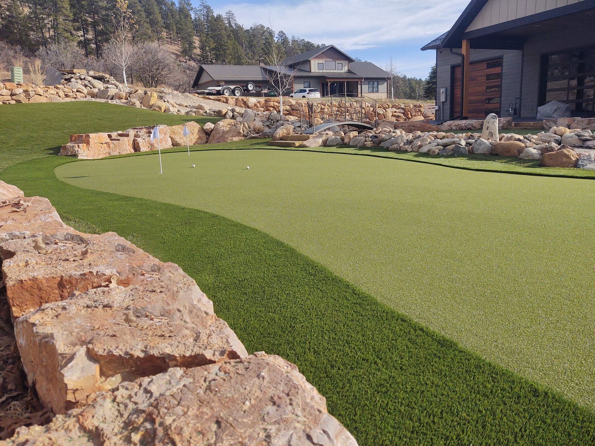 Golf green with artificial turf, bordered by stone walls and landscaping, with a house in the background