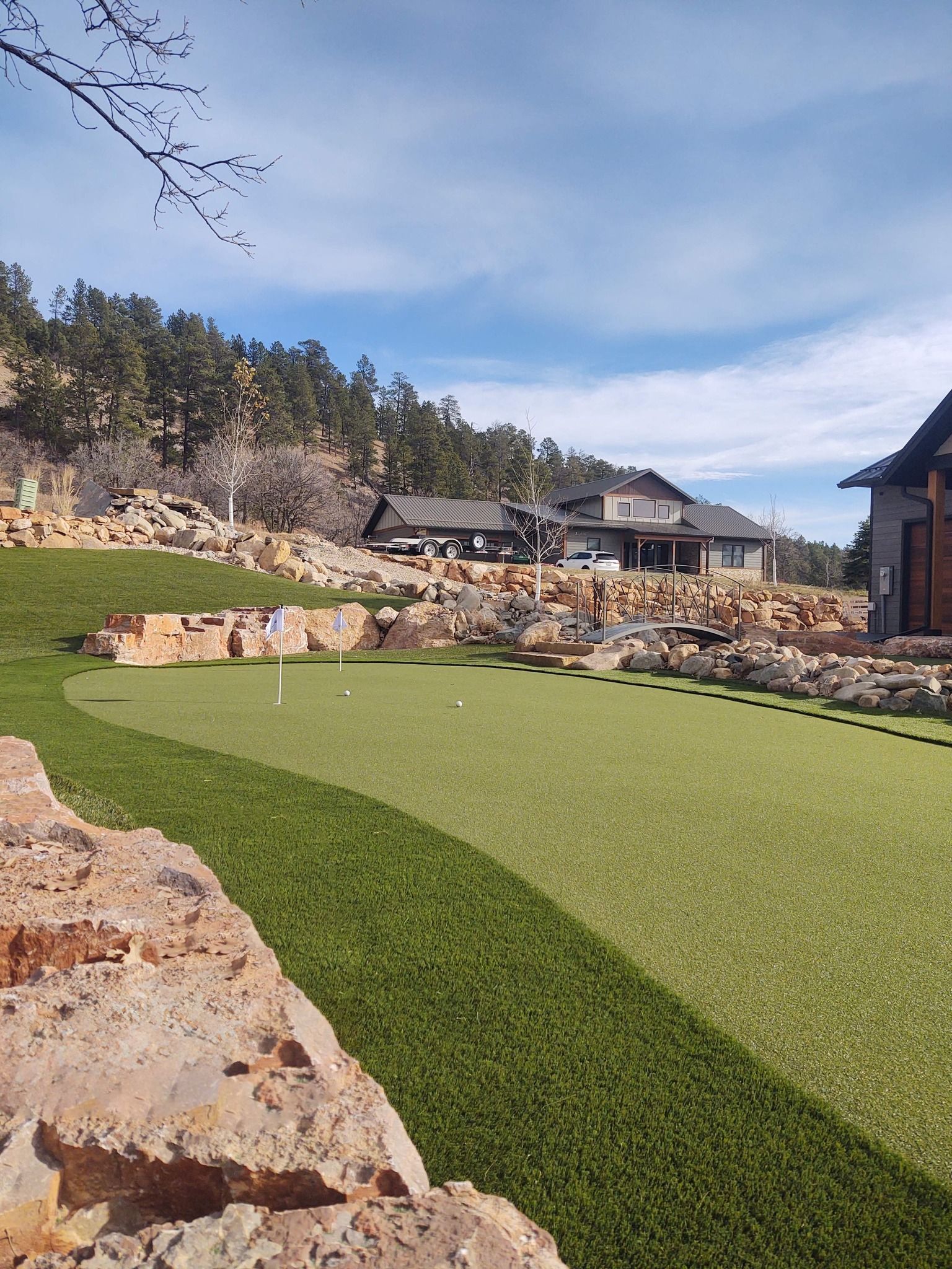 A backyard putting green with a luxury home in the background, surrounded by rock landscaping and trees, under a blue sky