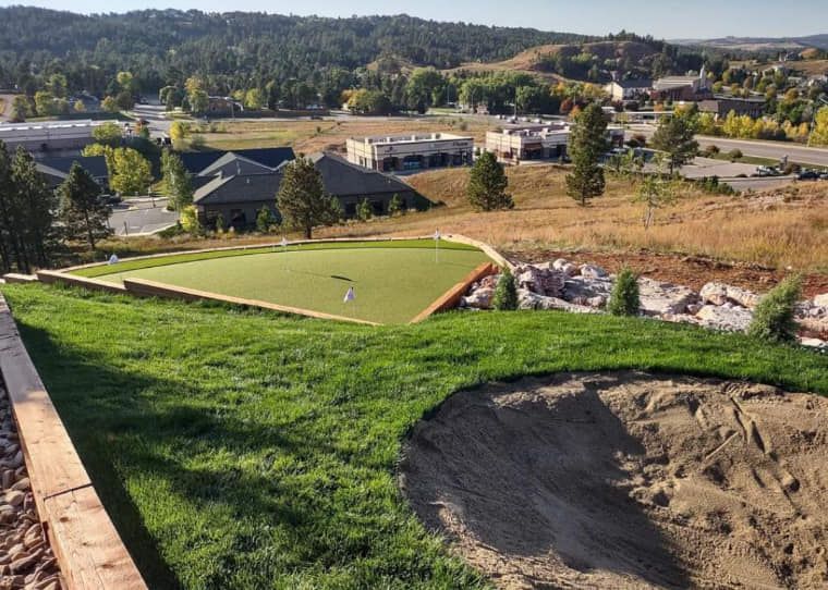Green golf putting area on a hill with a sand trap in the foreground, buildings, and trees in the background