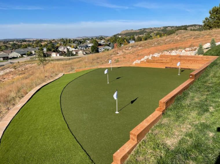 A backyard golf putting green with multiple holes, surrounded by landscaping and a view of a town
