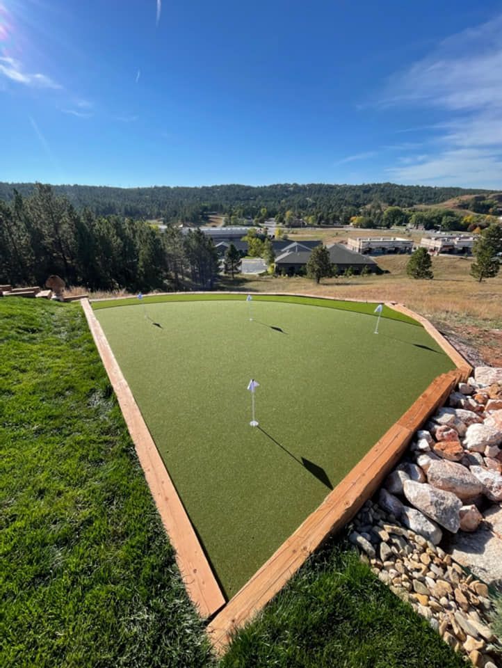 Green artificial turf putting green with multiple holes, bordered by wood and rocks, overlooking a valley
