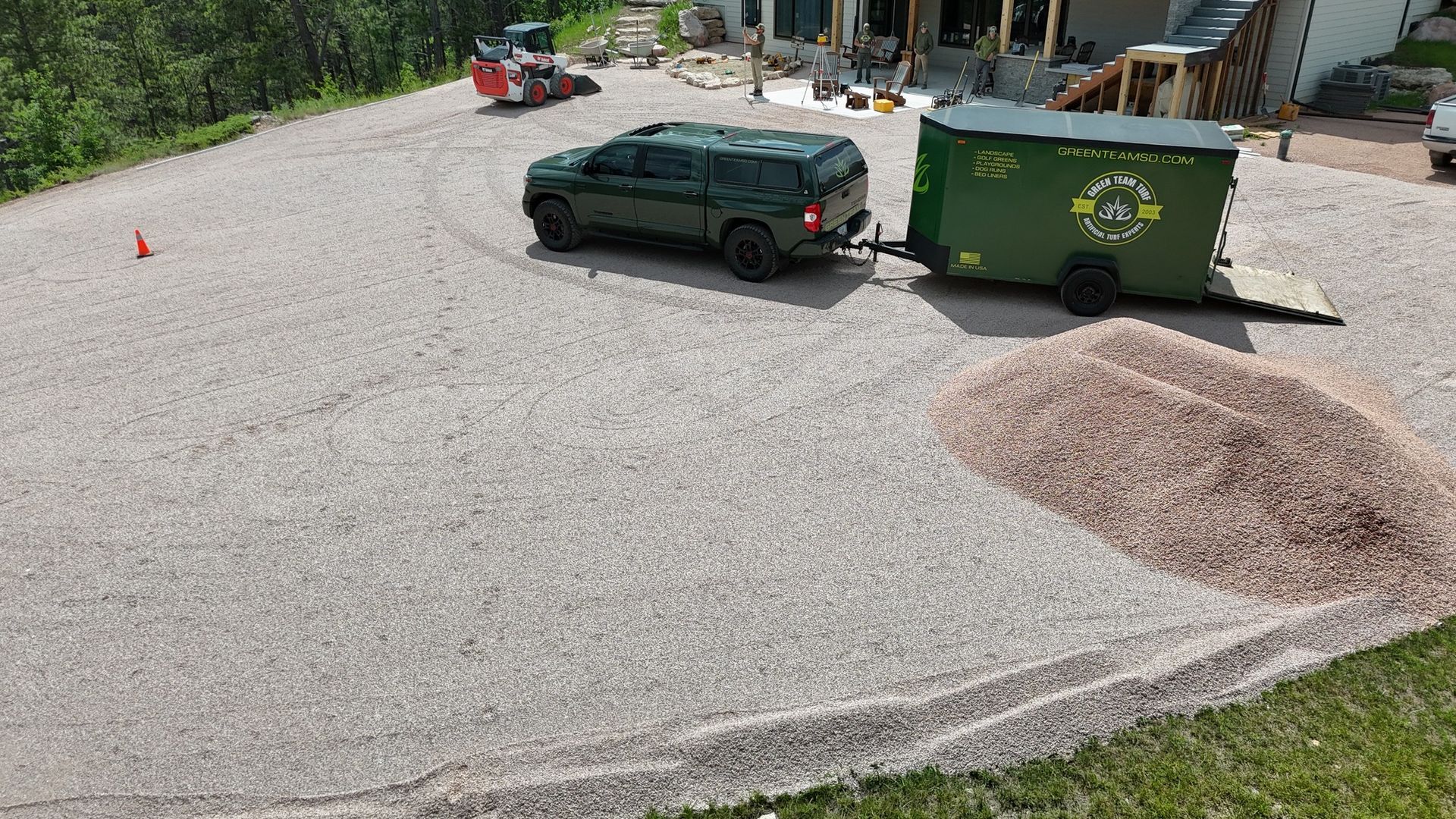 Green truck pulling a green trailer on a gravel driveway, with a pile of gravel to the right