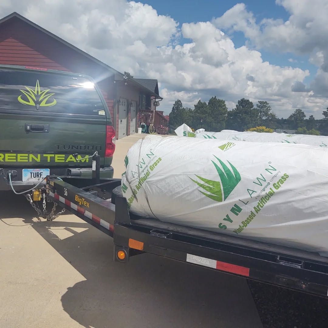 Truck towing a trailer loaded with rolled-up SynLawn artificial grass, parked near a building on a sunny day