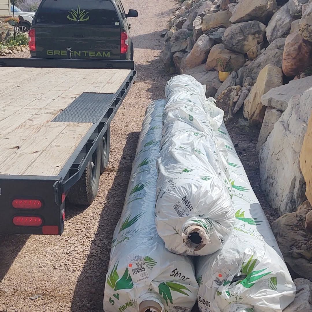 A truck with trailer next to rolls of landscaping fabric, near a rock retaining wall