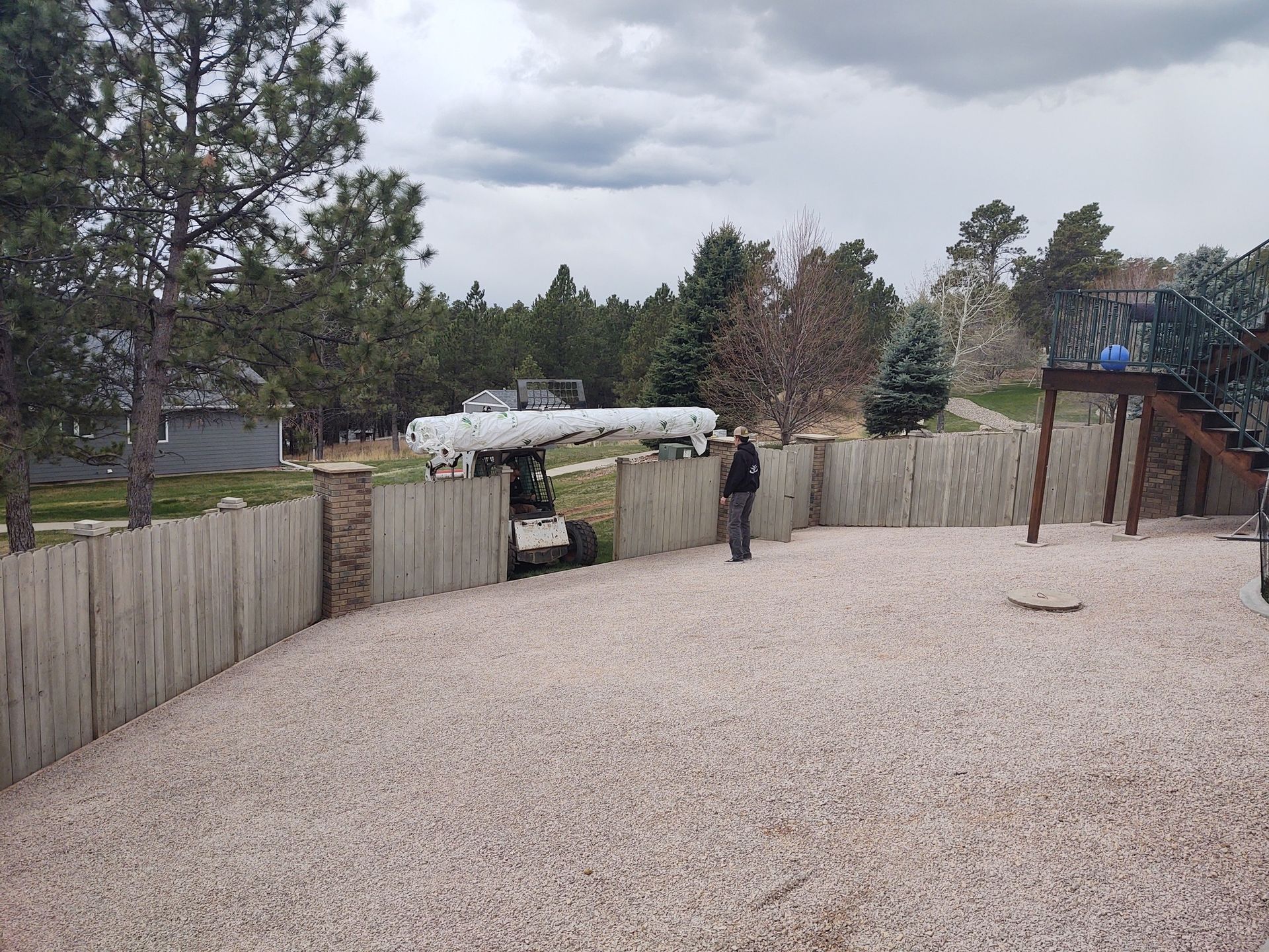 A person guides a small tractor carrying a large, white object through a gravel-covered yard surrounded by a wooden fence