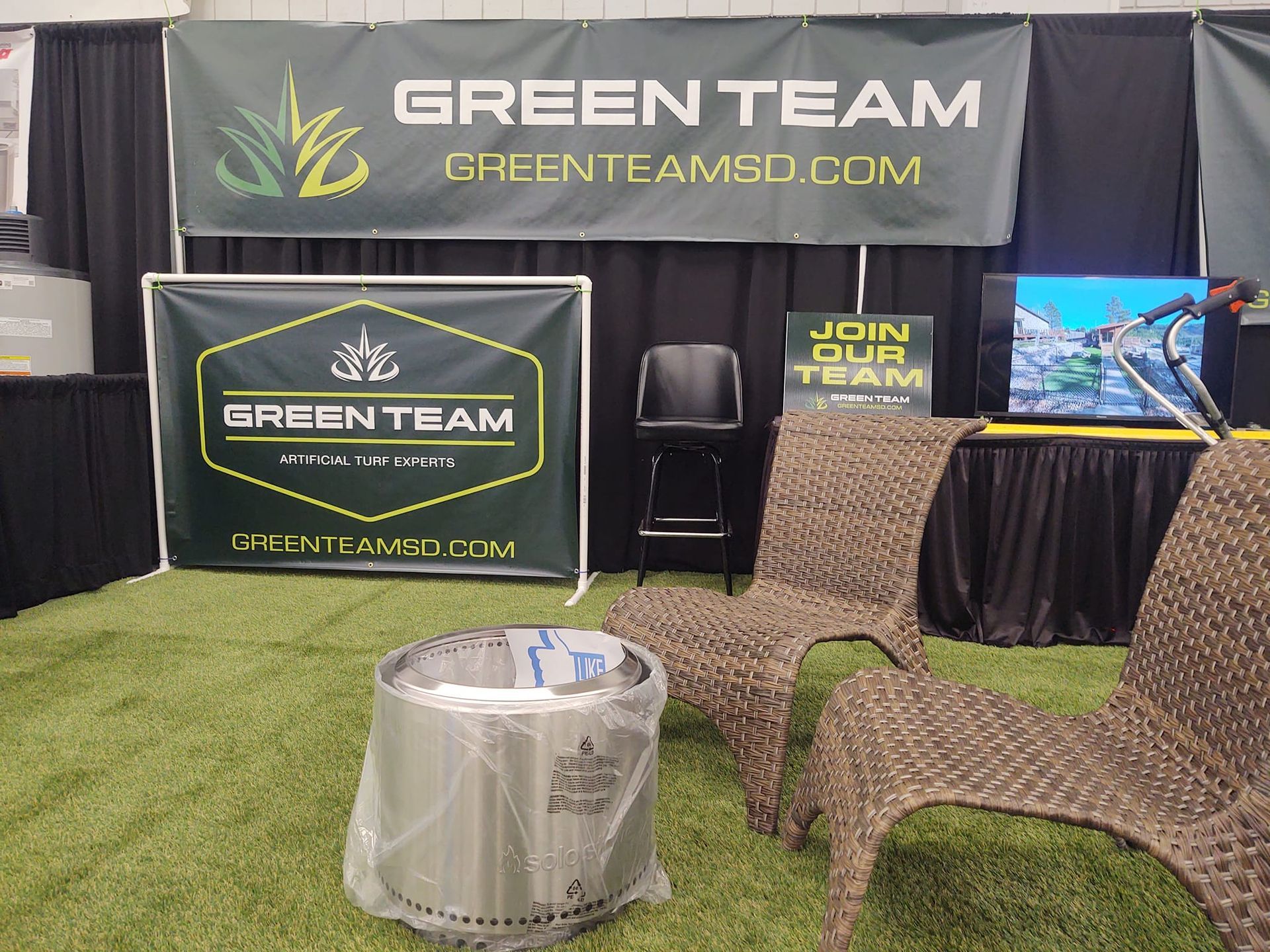 Green Team booth with signs, seating, a fire pit, and a TV at a trade show