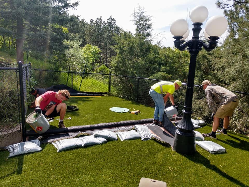 Three people installing artificial turf near a fence and a lamppost, pouring materials