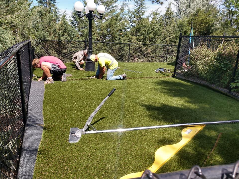 Three workers installing artificial turf in a fenced yard, using hand tools