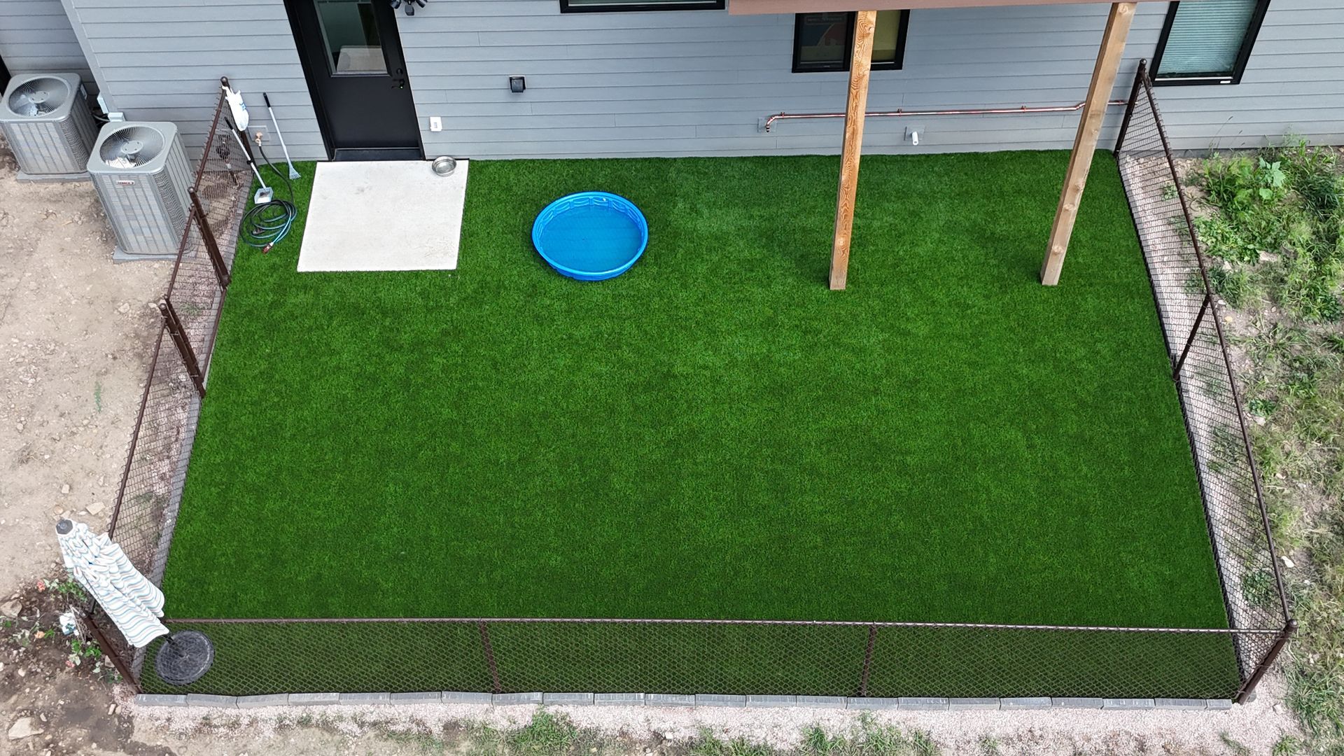 Overhead view of a fenced yard with green turf, a blue dog pool, and a building with a black door.