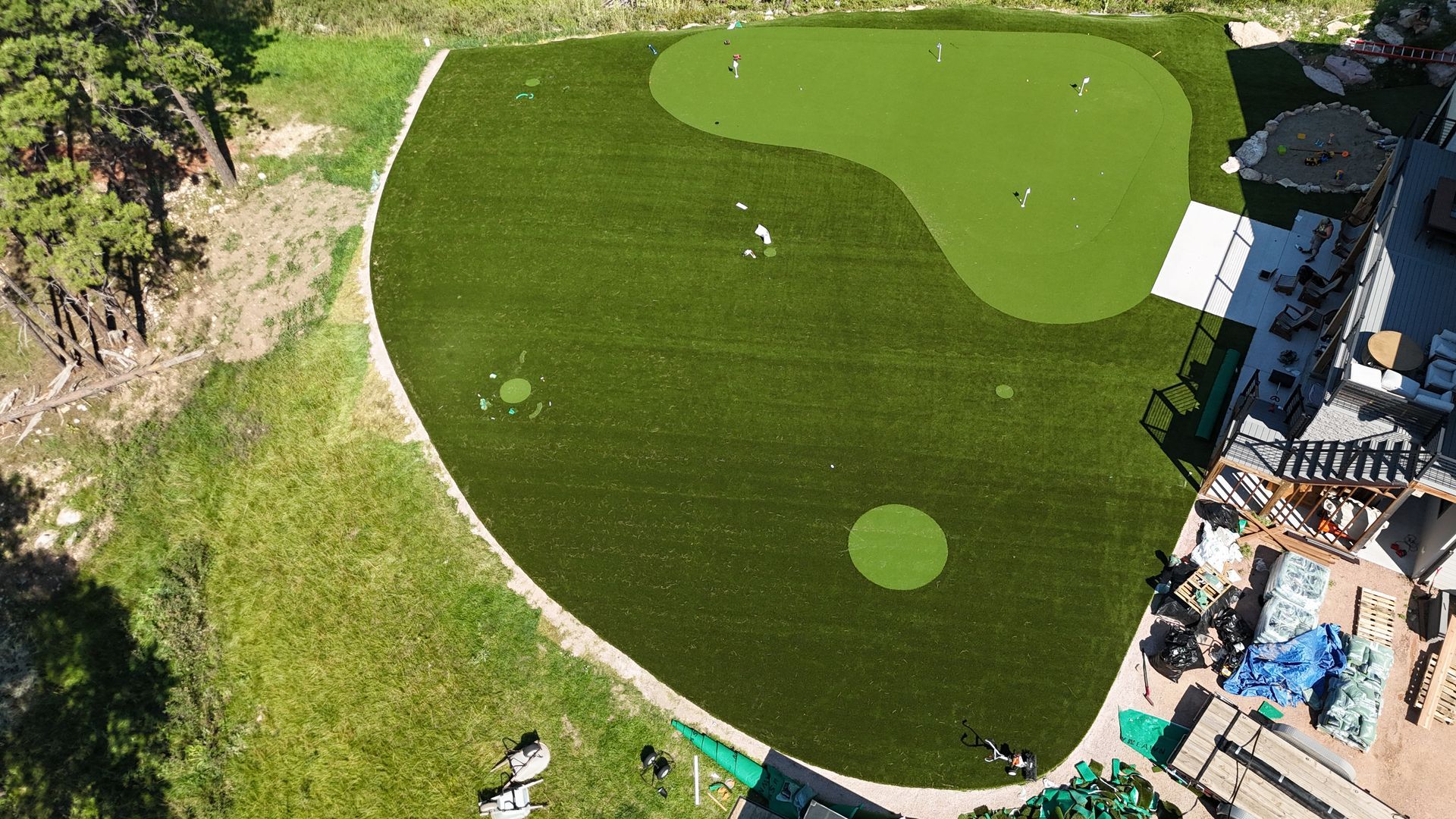 Aerial view of a green golf putting area with players, on a grassy hillside next to a building.