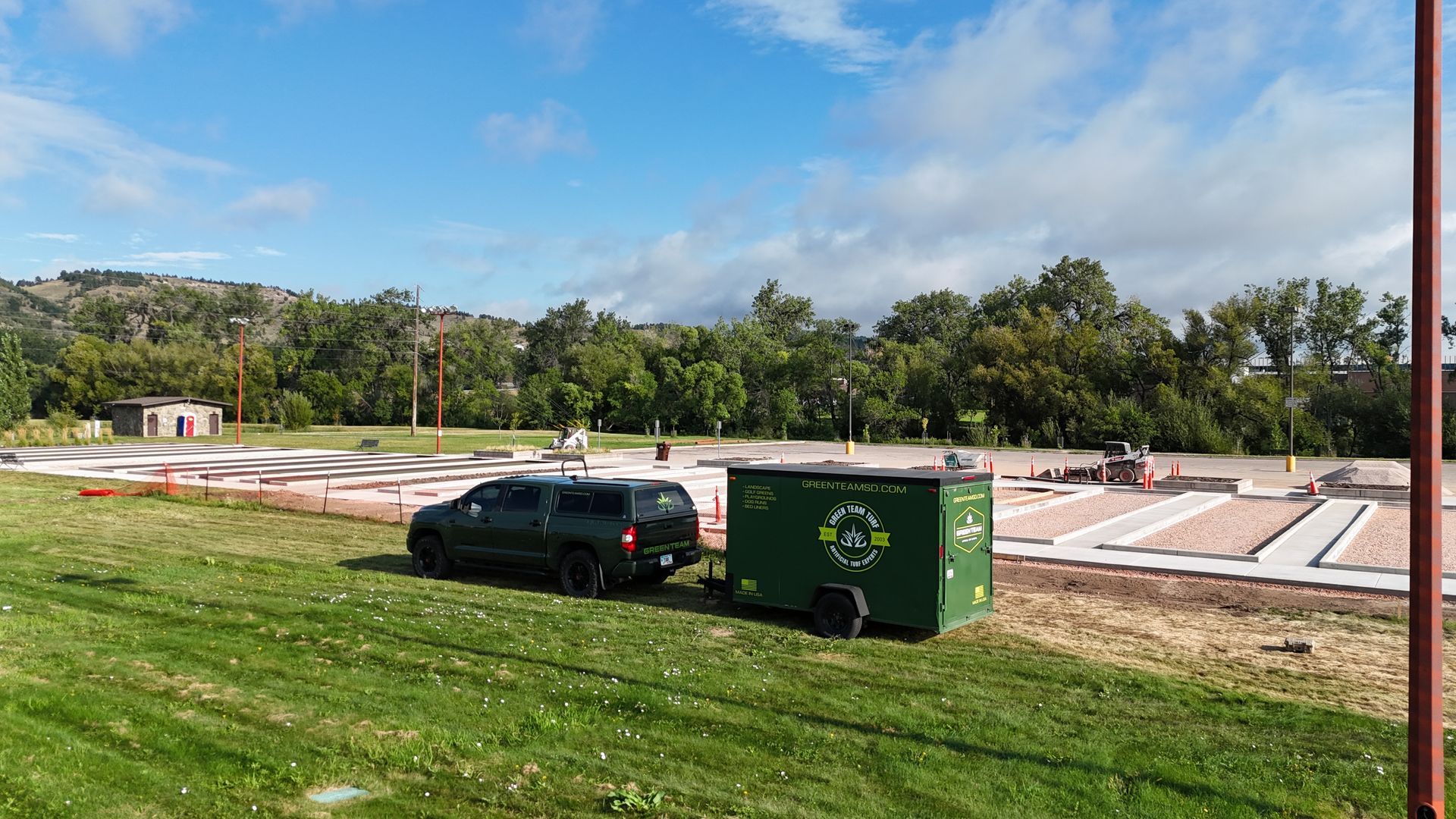 Black truck towing a green trailer on a grassy area near a construction site under a blue sky.
