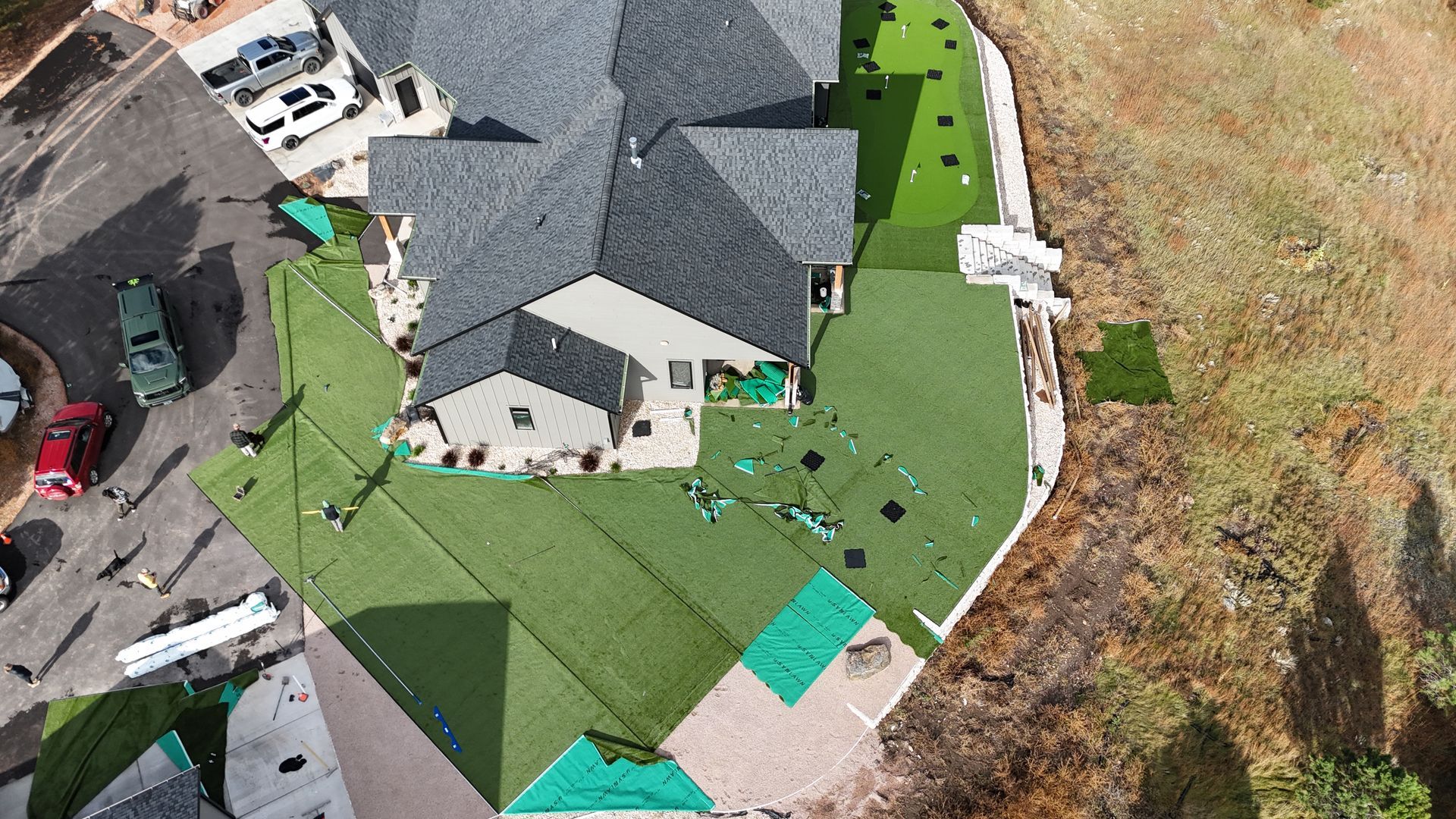 Aerial view of a house with dark roof and green turf lawn, showing installation with green netting and a nearby parking area.