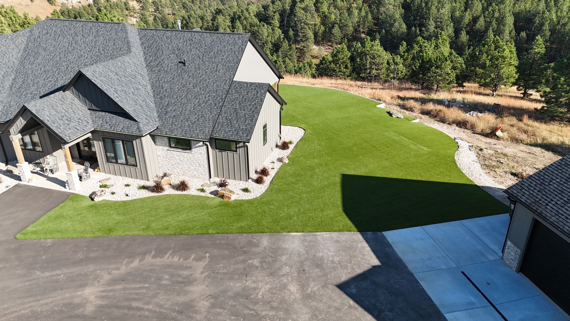 Gray house with dark roof and green lawn, with driveway and trees in the background.