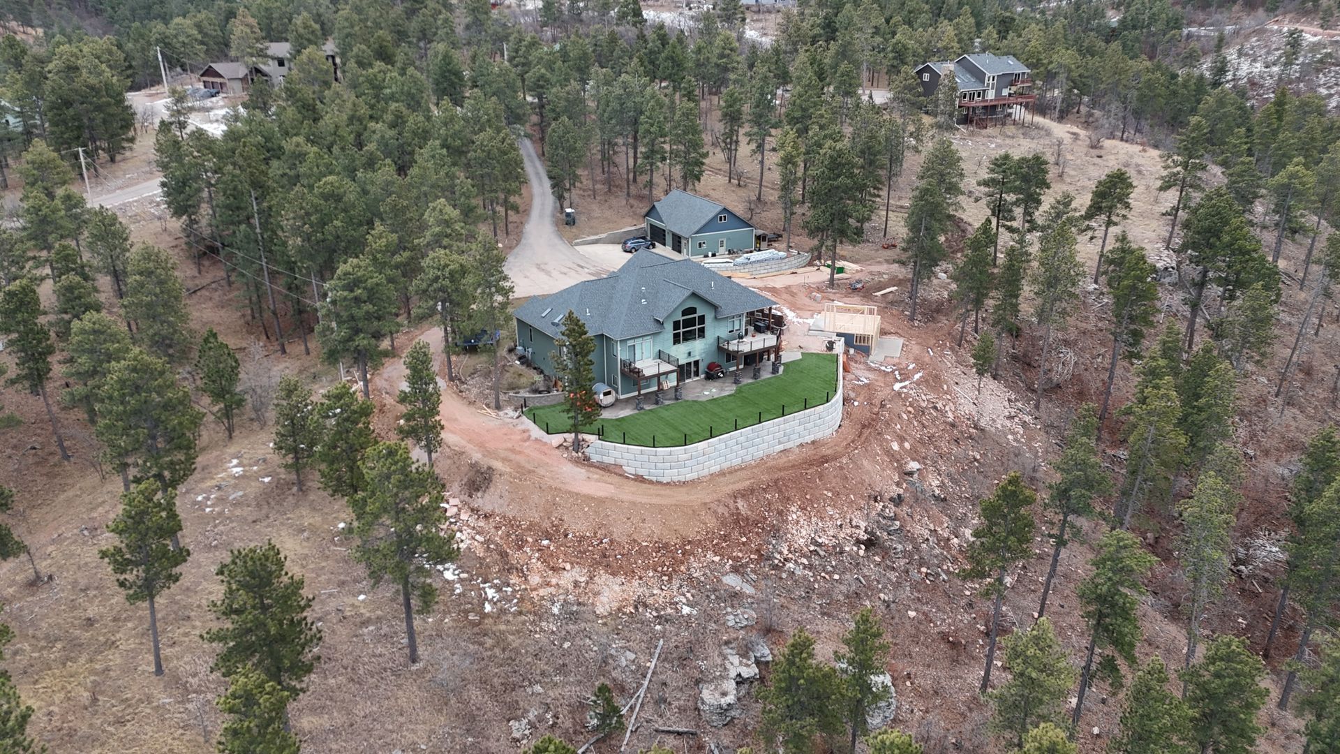 Aerial view of a house nestled in a forest on a hillside with a retaining wall and trees.