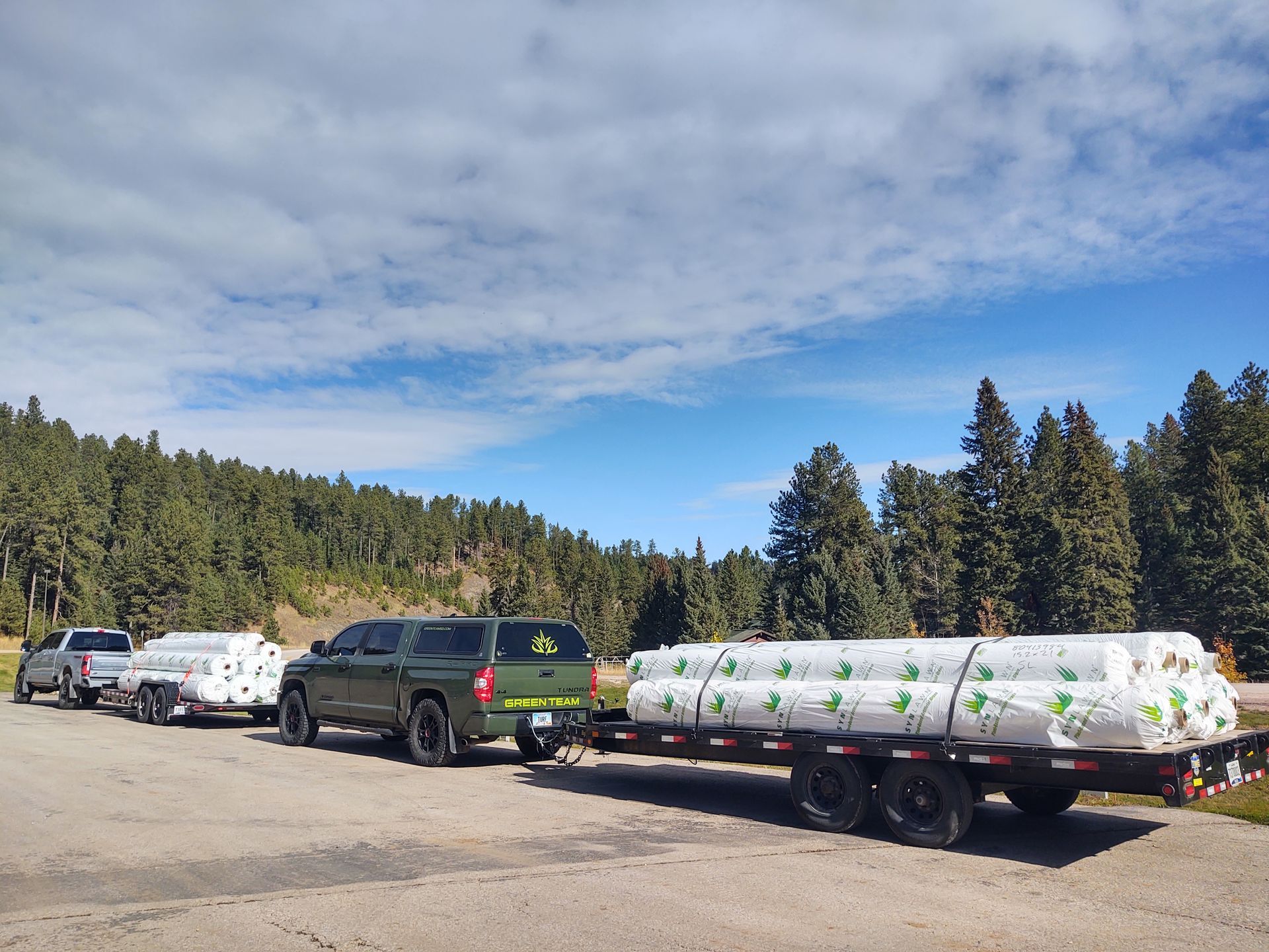 Vehicles hauling cargo, parked in a lot. Green truck towing a trailer with rolled items, trees and blue sky background.