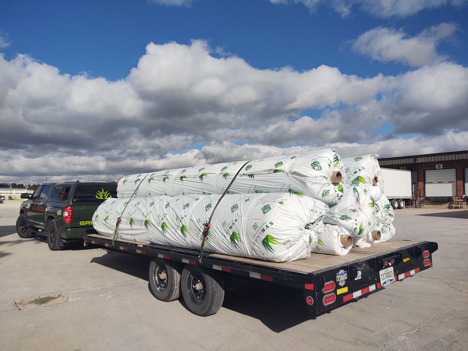 Truck pulling a trailer loaded with several large rolls of white plastic-wrapped materials, under a cloudy blue sky.