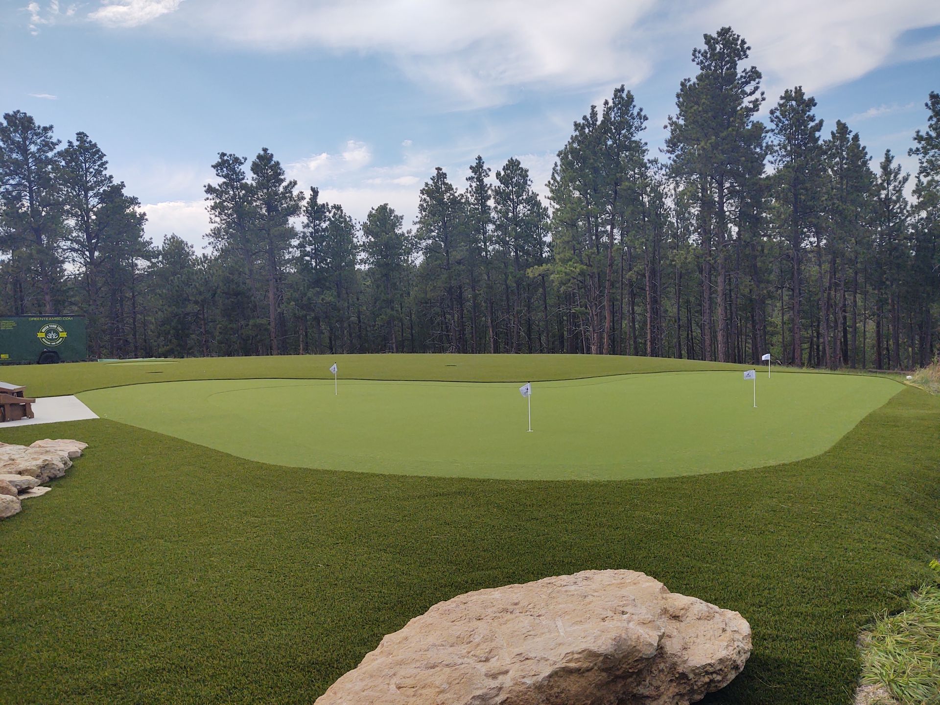 Green putting area with three flags, surrounded by artificial turf and trees. Blue sky.