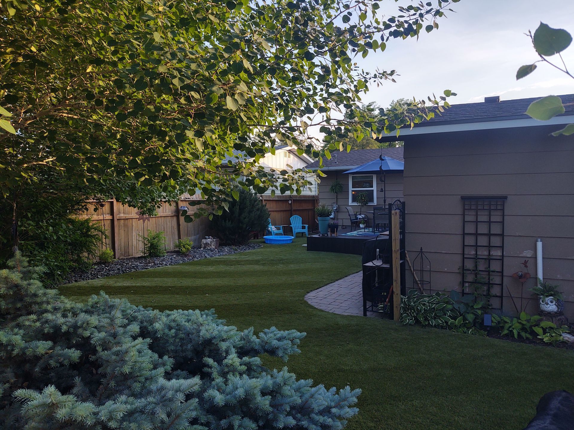 Backyard with green lawn, trees, brown house, blue chairs on deck, sunny.