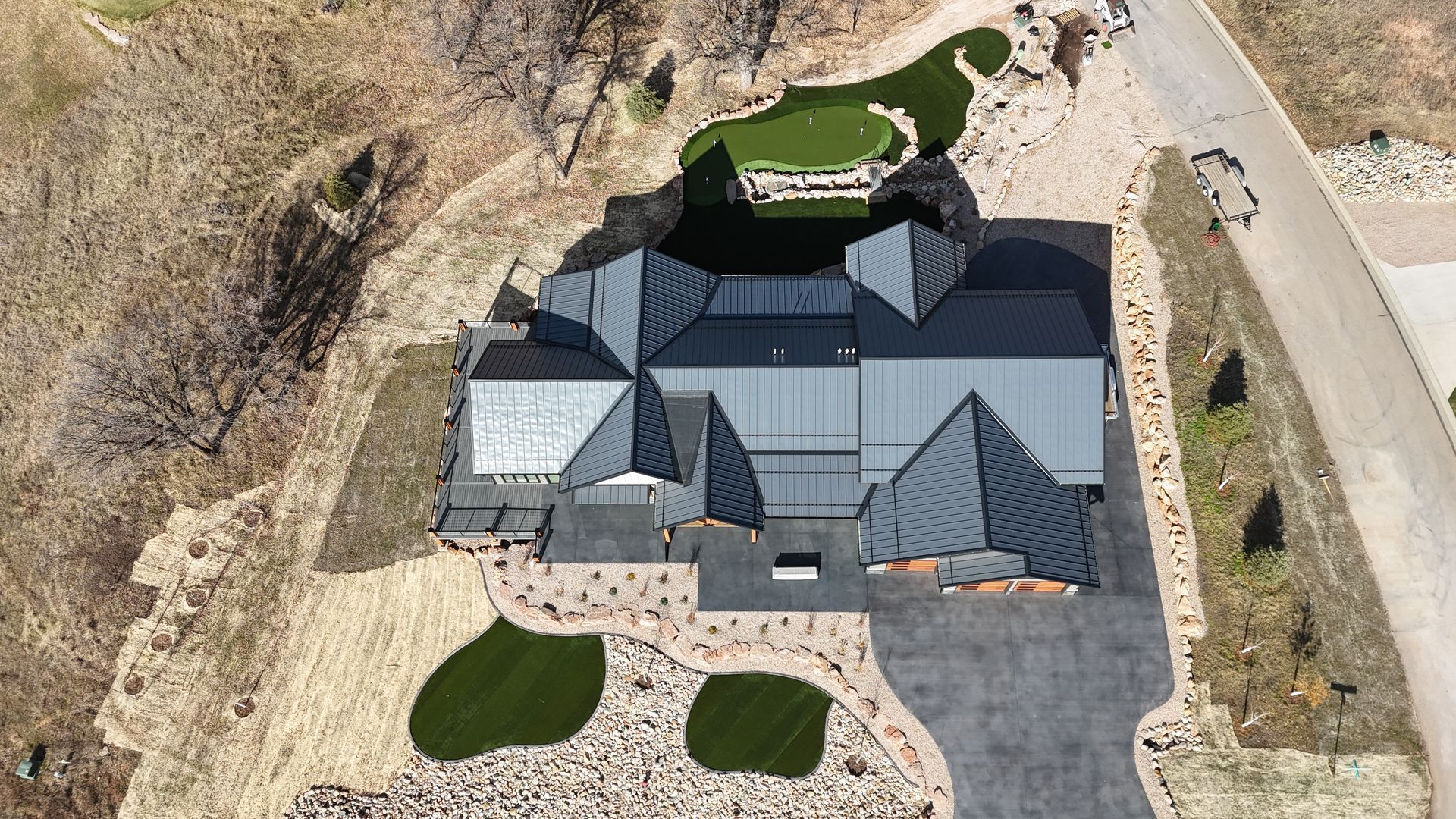Aerial view of a modern house with a complex, dark gray roof, surrounded by landscaping, a driveway, and barren hillside.