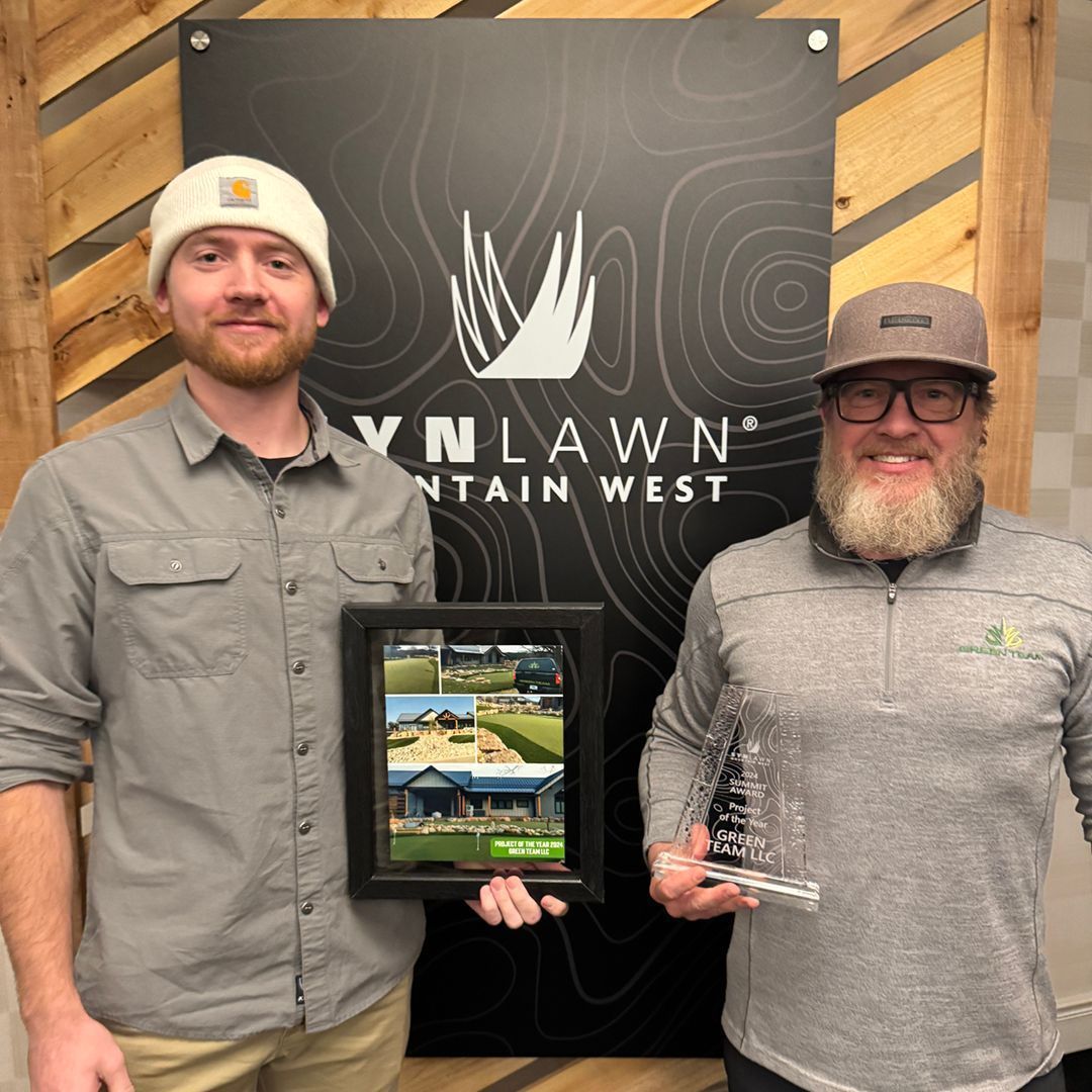 Two men holding awards in front of a company logo, smiling. The backdrop has wood paneling.
