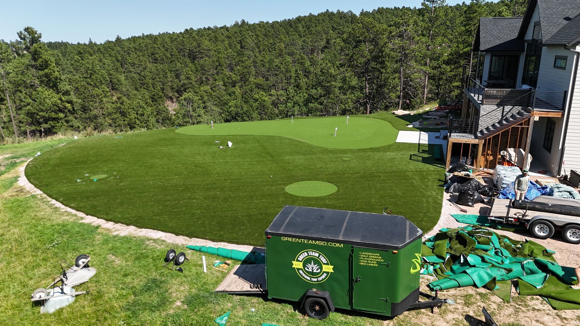 Artificial turf golf green installation at a home. Green turf, trailer, trees, and house.