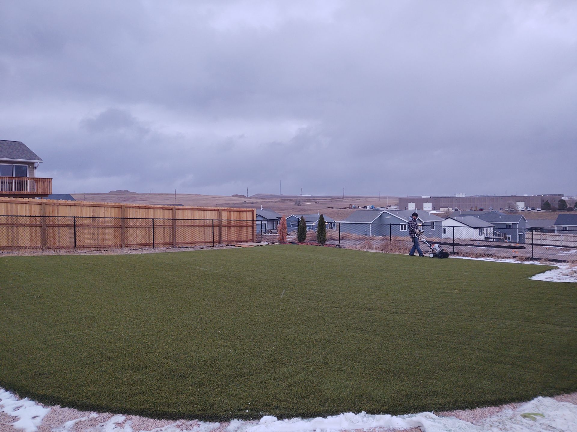 Backyard with green artificial turf, fence, and a person near equipment under an overcast sky.