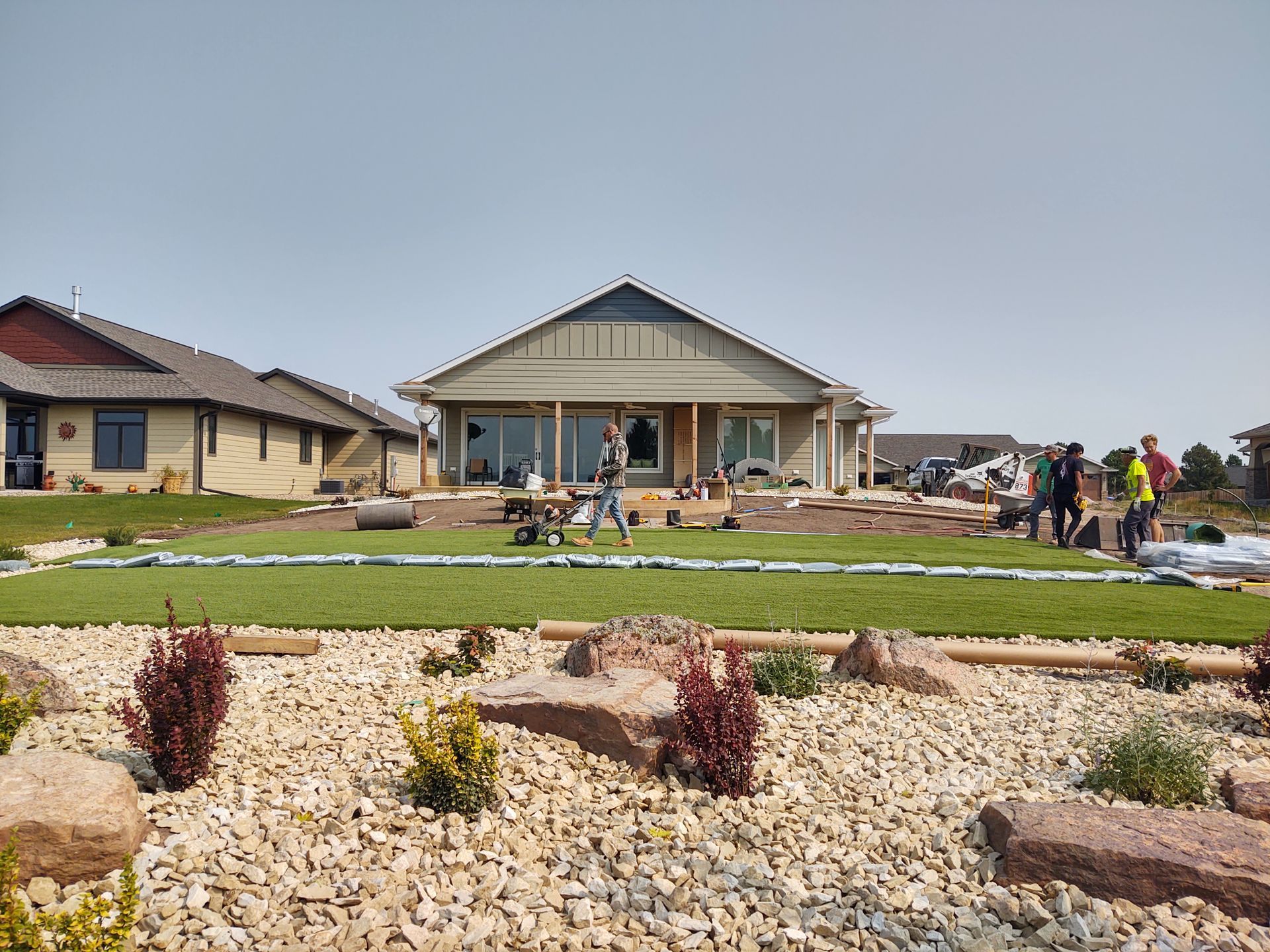 Landscaping in progress in front of a house. Workers install green lawn; large rocks and bushes in foreground.