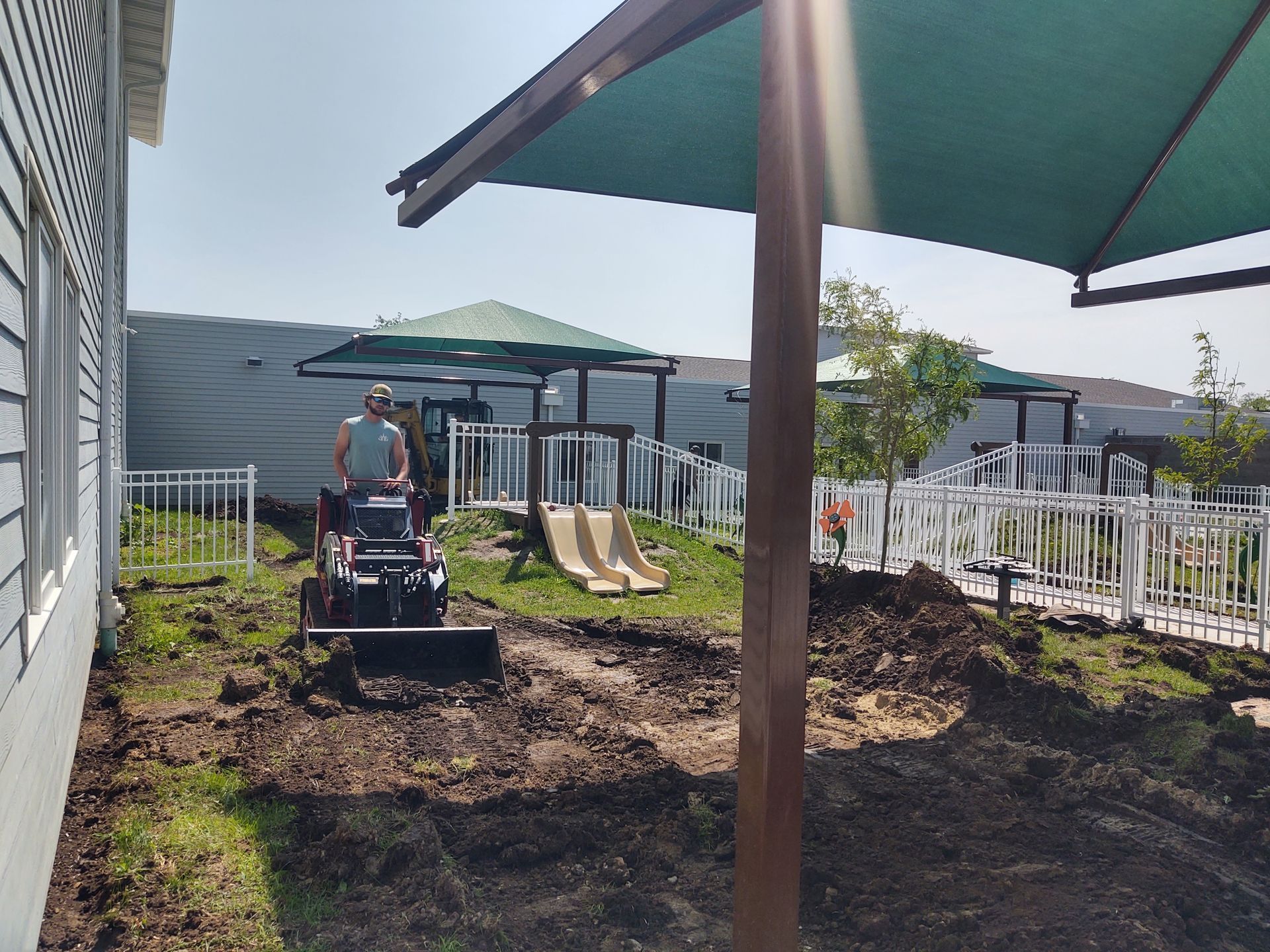 Man operating a small tractor on a dirt area next to a building with a playground in the background.