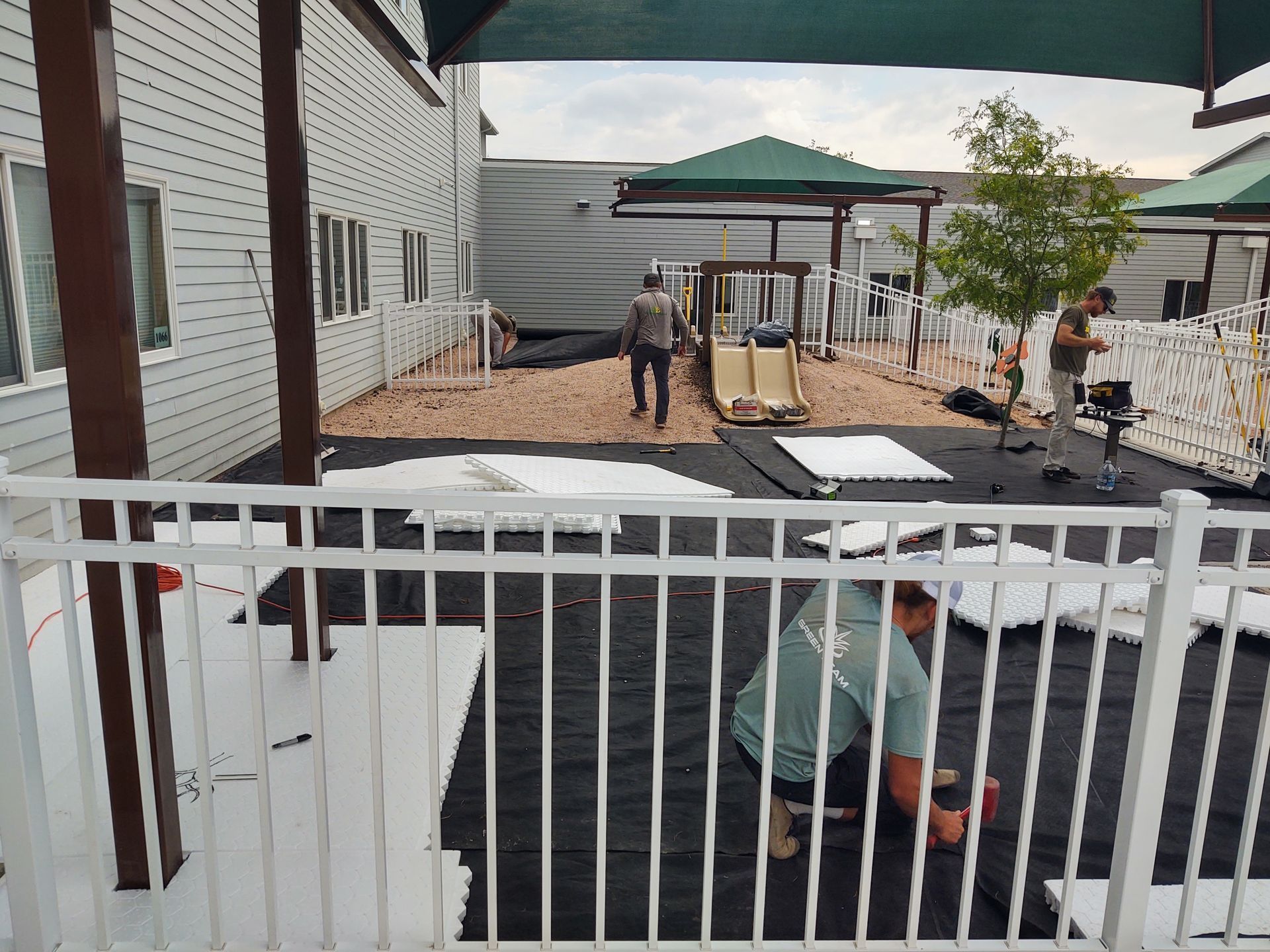 Workers installing white fence panels and ground cover in an outdoor play area.