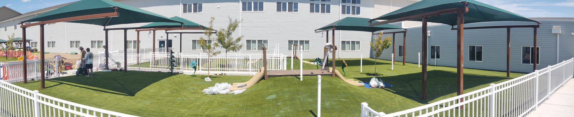 Outdoor play area with green grass, white fence, and covered structures in front of a white building.