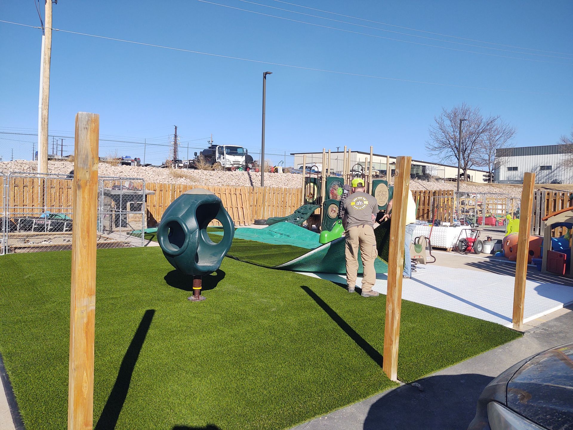 Playground with green turf, equipment, wooden posts, and a person adjusting a shade.