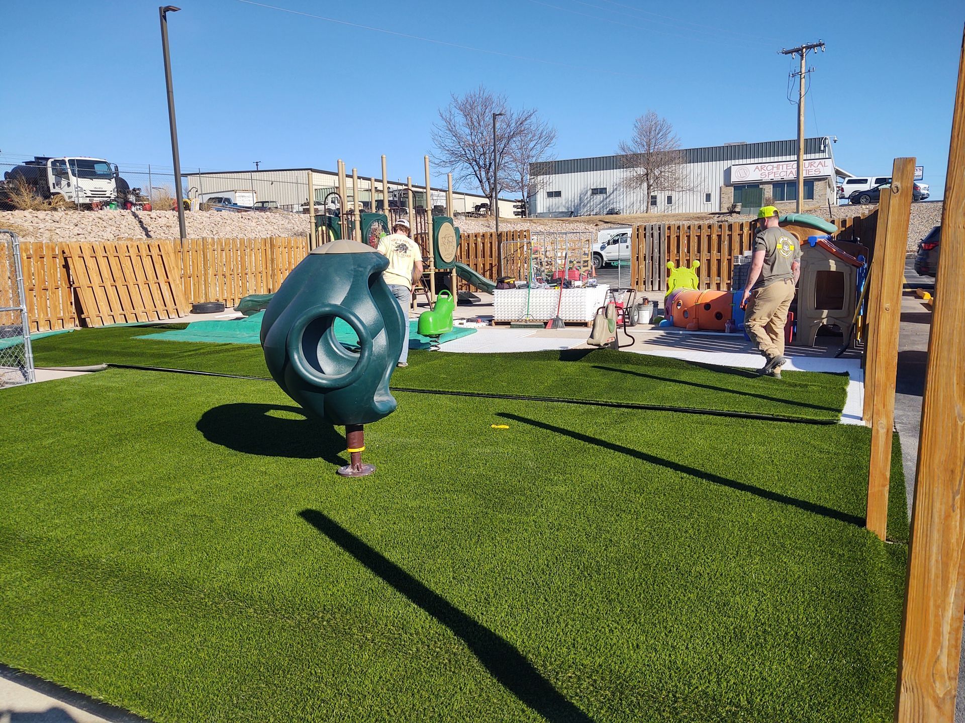 A playground with artificial turf, various play structures, and a wooden fence. Two people are working near the fence.