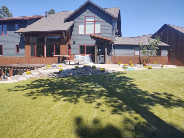 Backyard view of a gray house with red-framed windows, a grassy lawn, and a tree casting a shadow.