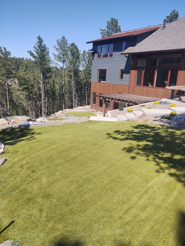 Green lawn slopes toward a two-story home with a dark-roofed pergola, surrounded by trees on a sunny day.