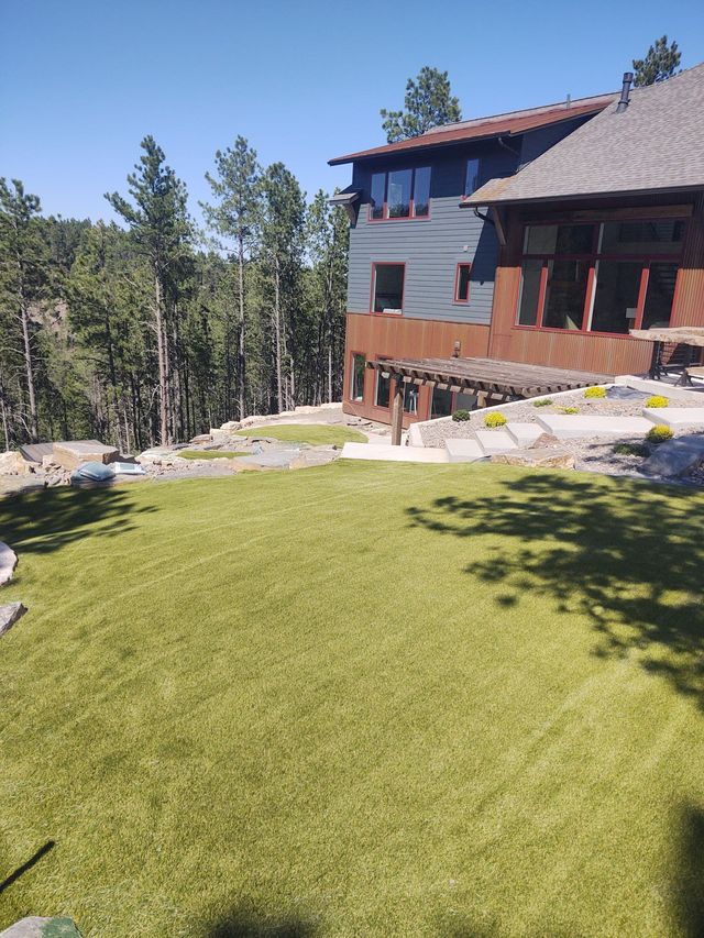 Green lawn slopes toward a two-story home with a dark-roofed pergola, surrounded by trees on a sunny day.