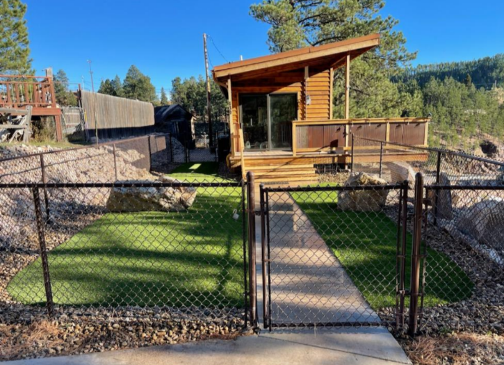 Small wooden cabin with a deck, artificial turf, and chain-link fence.