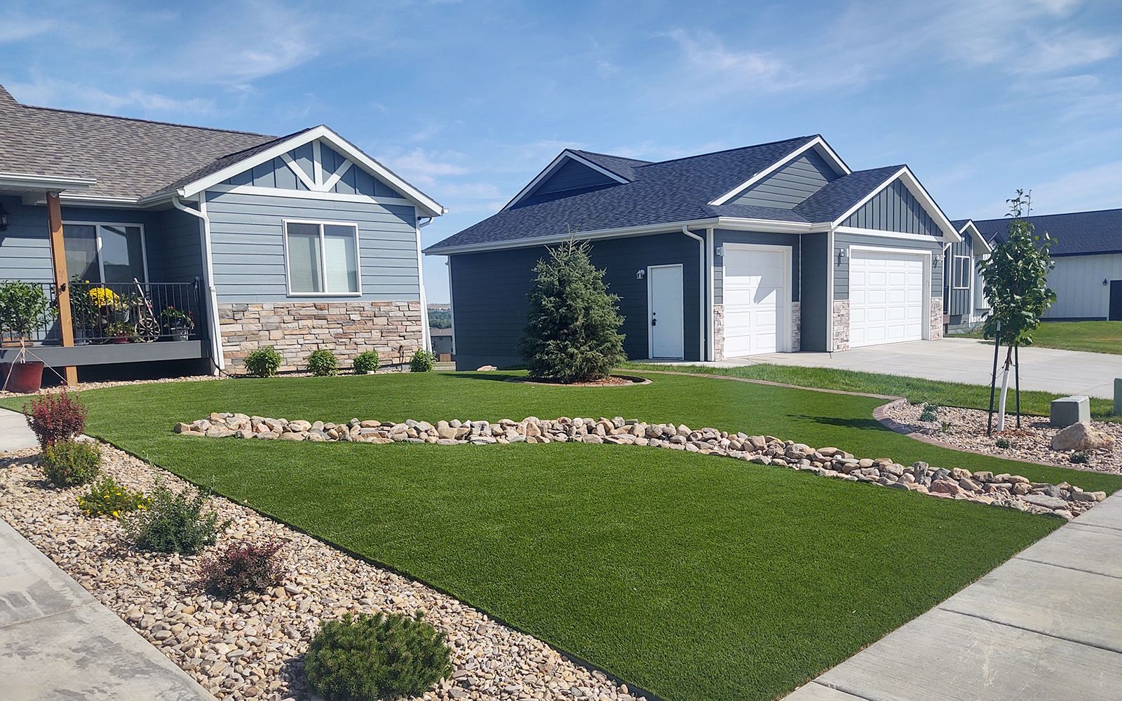 Two suburban houses with manicured lawns, rock beds, and blue siding on a sunny day.