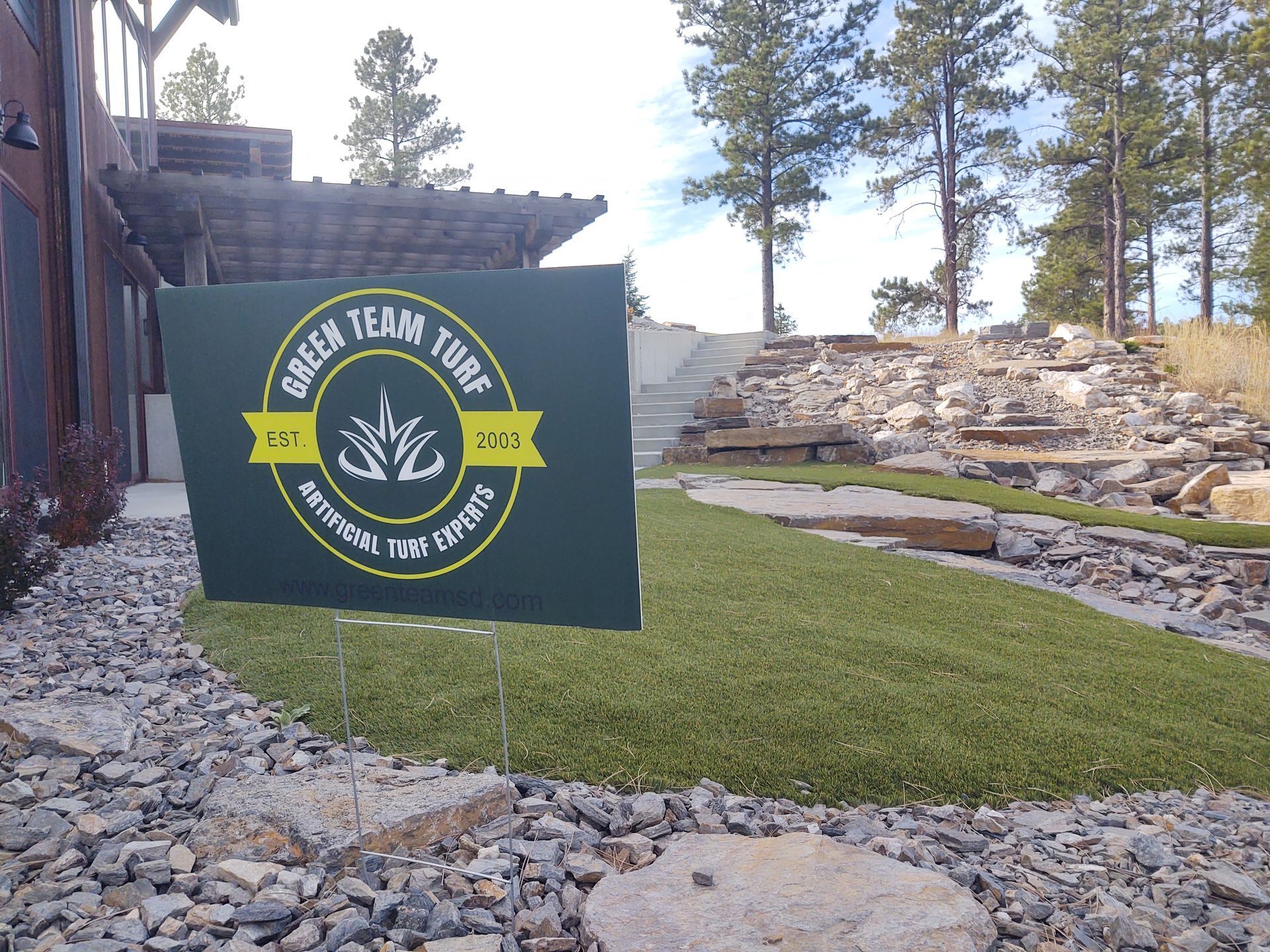 Green Team Turf sign in front of artificial turf display, with a rocky landscaping and trees in the background.
