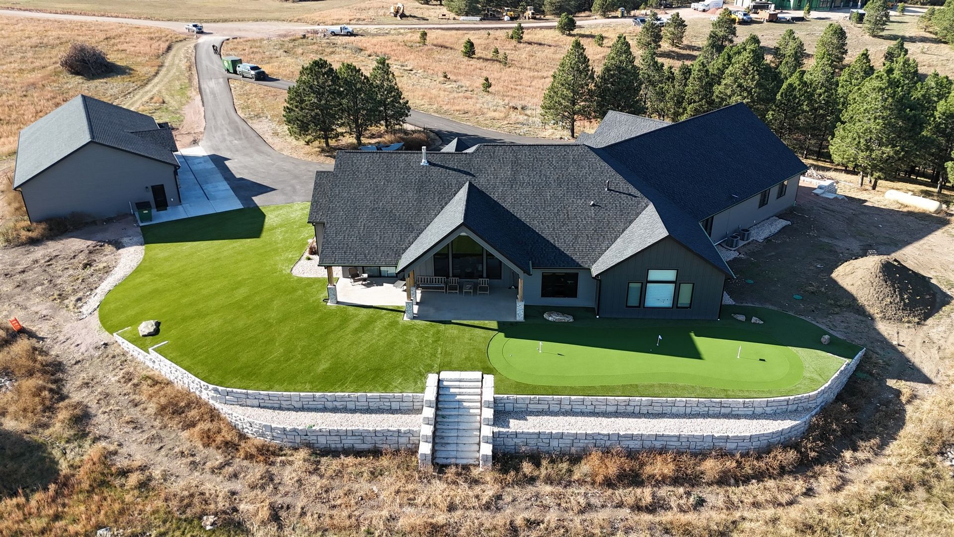 An aerial view of a dark-gray home with a putting green, stone retaining walls, and a separate garage on a grassy lot.