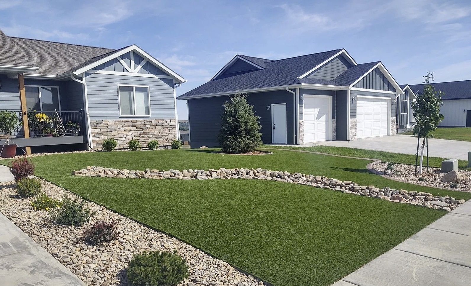 Two suburban houses with blue siding, stone bases, and front lawns with rock landscaping features under a sunny sky.