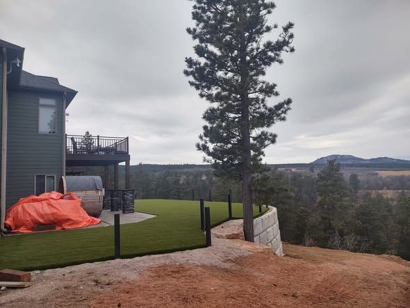 House with green siding and a deck, next to a newly fenced artificial lawn with a view of hills