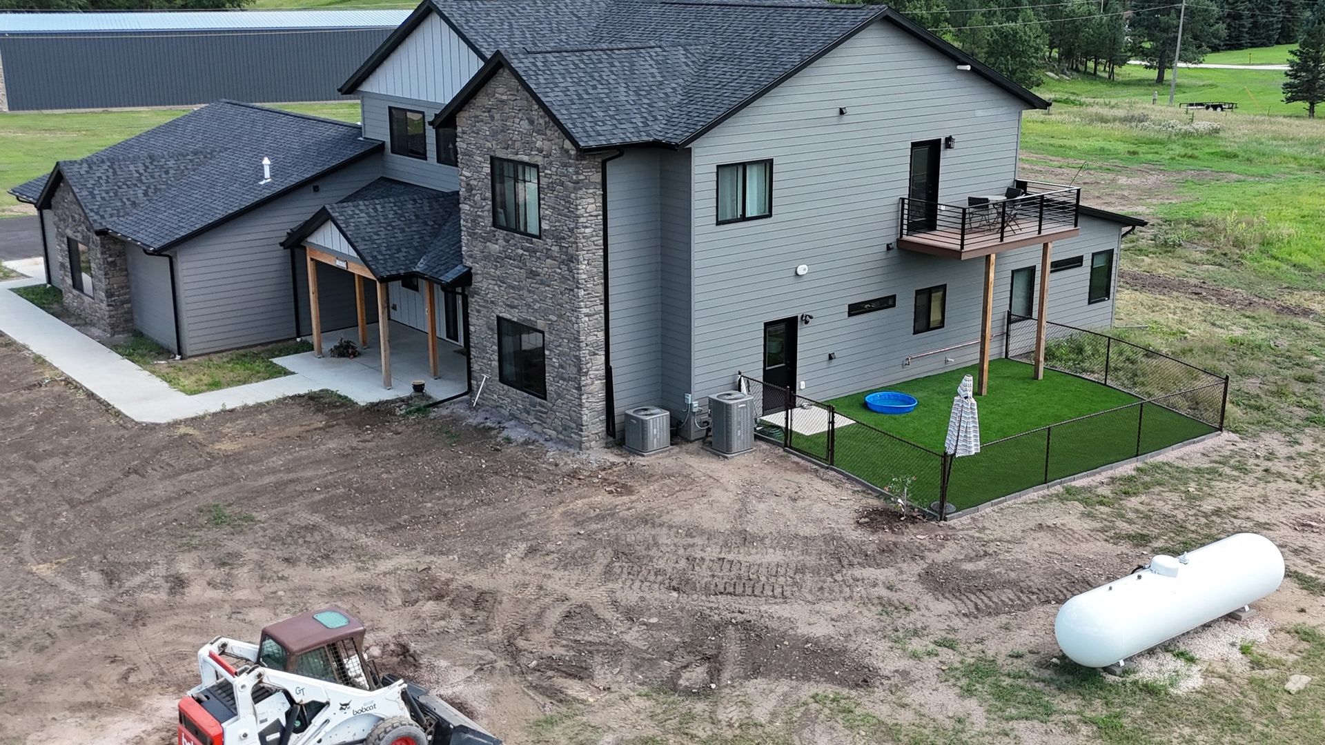 Two-story house with gray siding, stone accents, and a small fenced yard; a propane tank and construction equipment are in the foreground