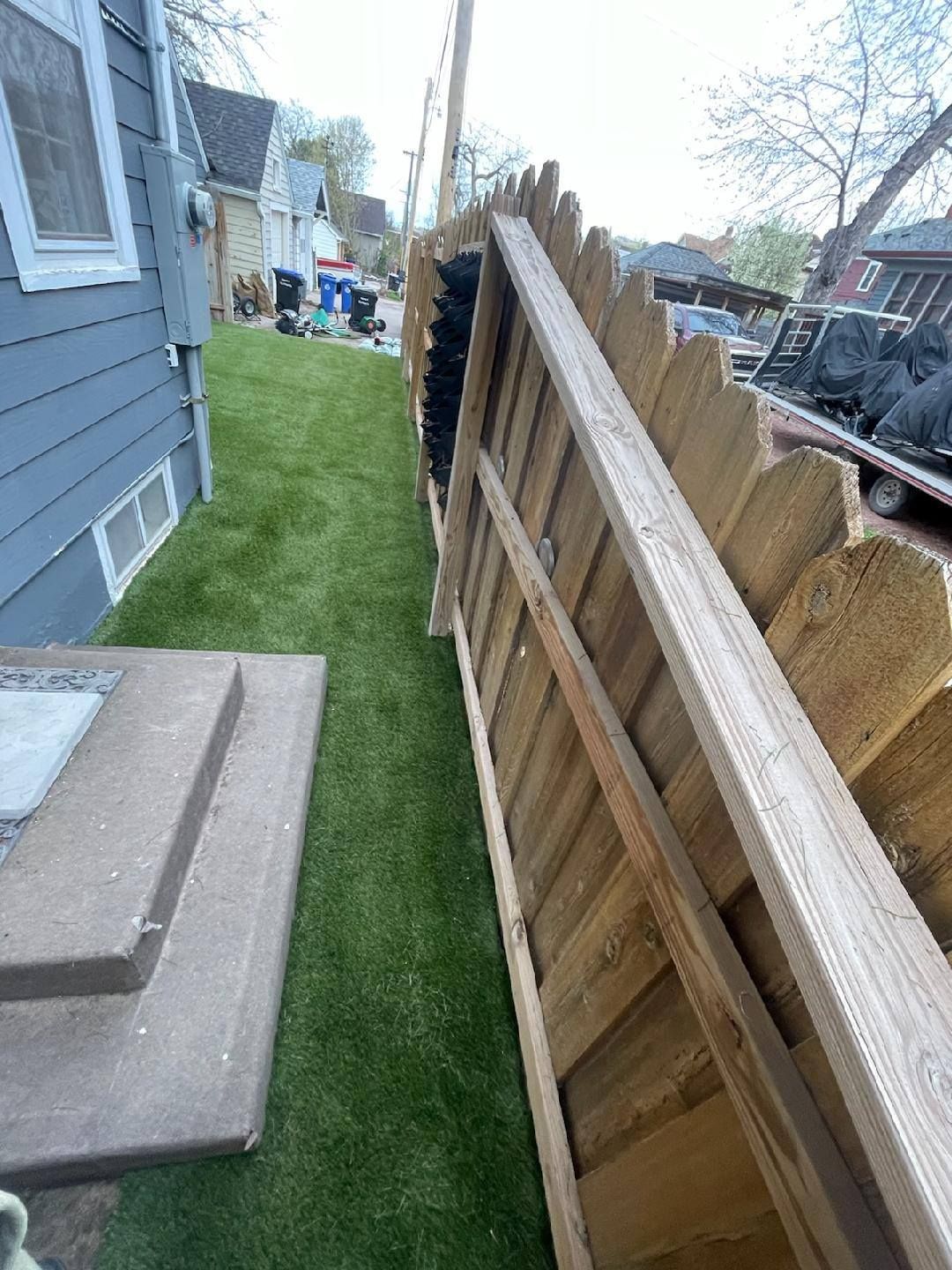 A wooden fence leaning against an artificial turf lawn next to a house with blue siding