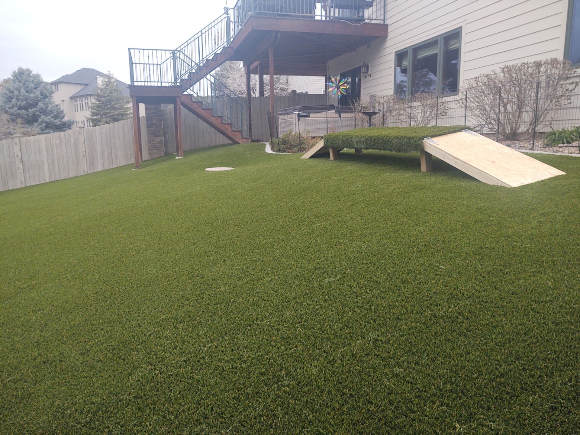 A backyard with artificial green grass, wooden deck, stairs, and a cornhole game set