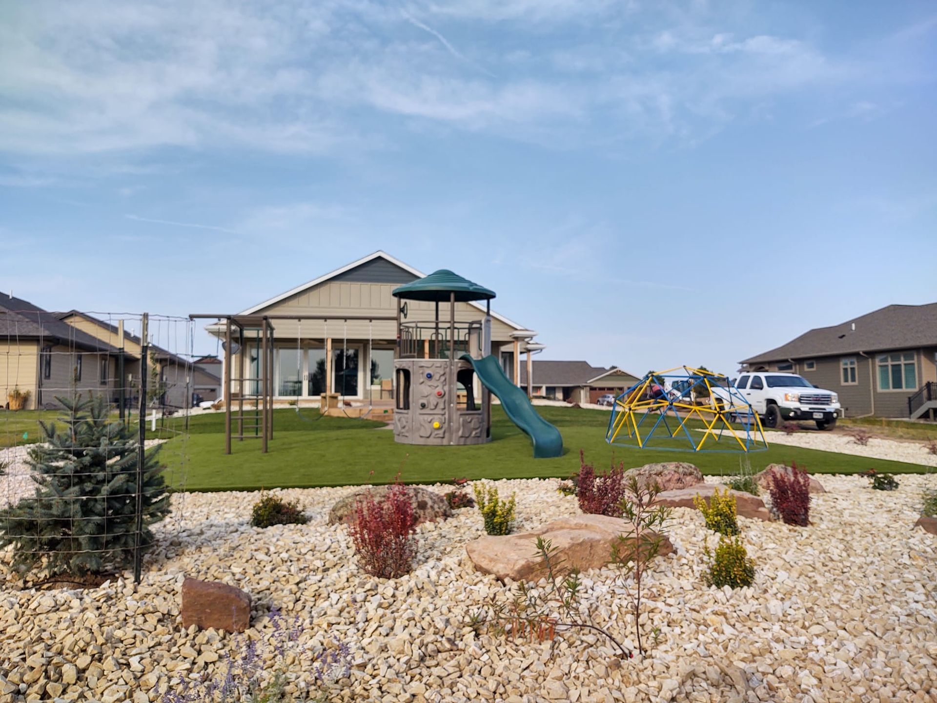 A backyard playground with a climbing tower, slide, and swing set on artificial turf, surrounded by white landscaping rocks.