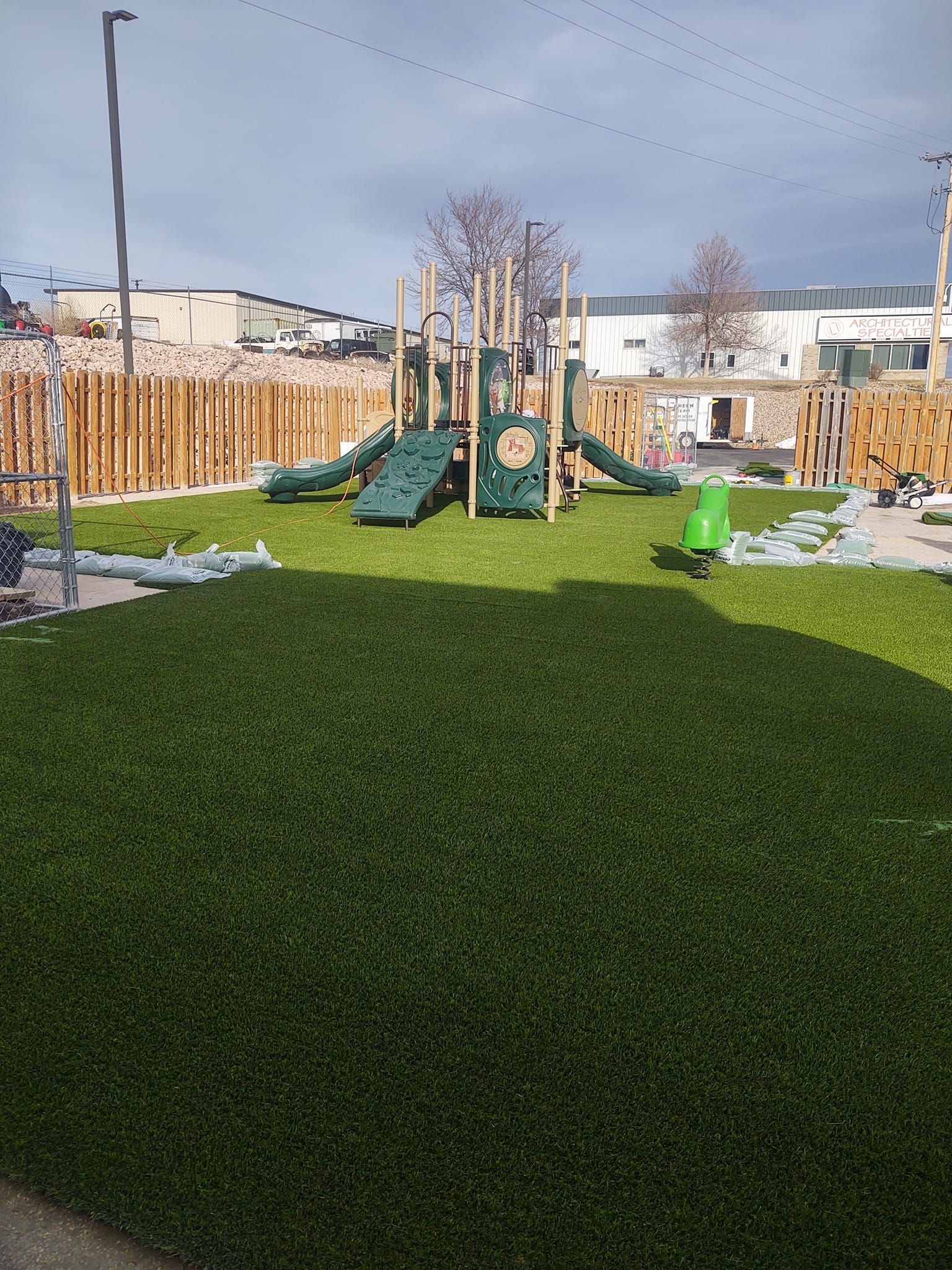 Playground with green turf, slides, climbing structures, and wooden fence on a sunny day
