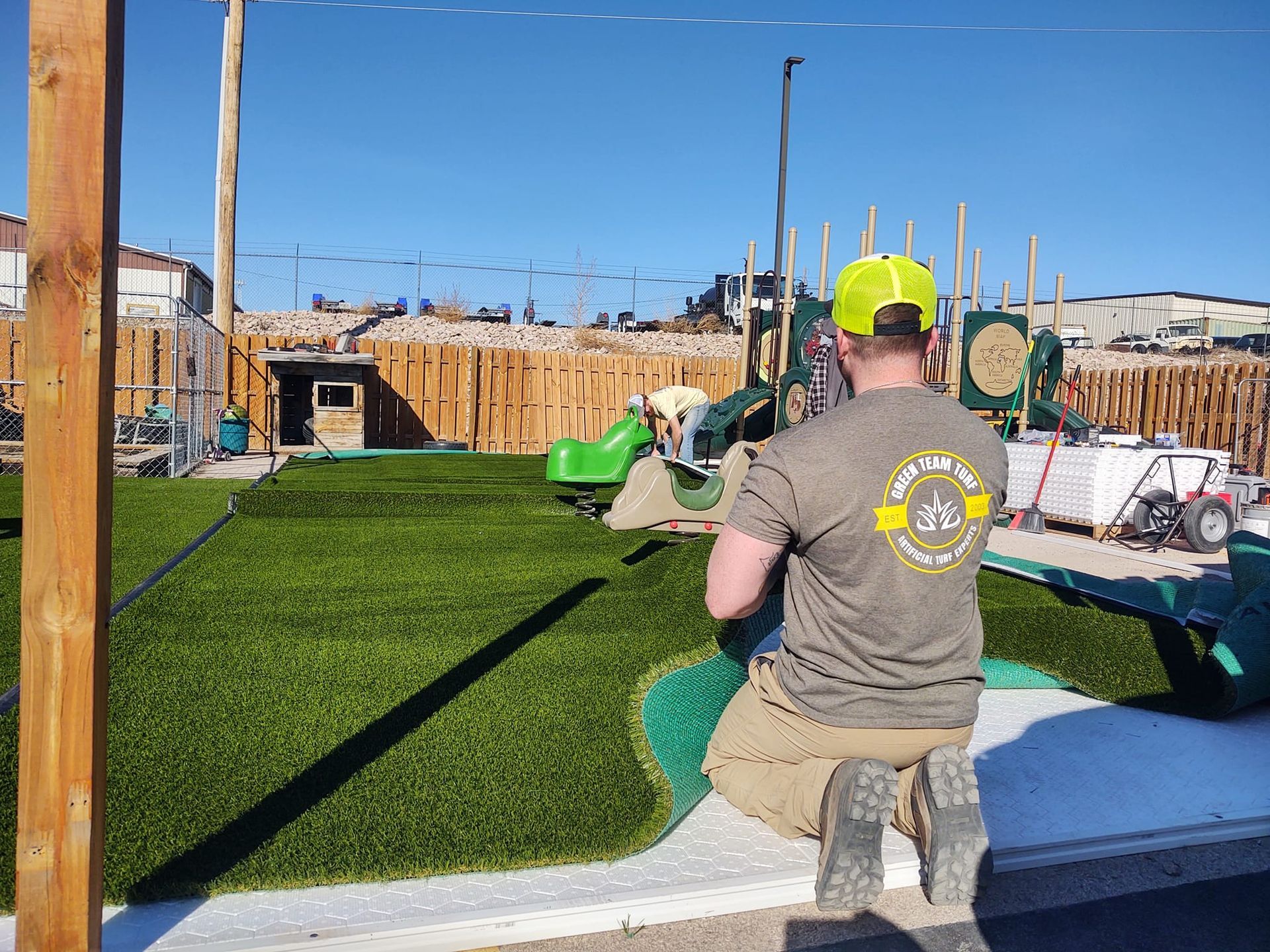 Person installing artificial turf in a playground area with a slide, under a sunny sky