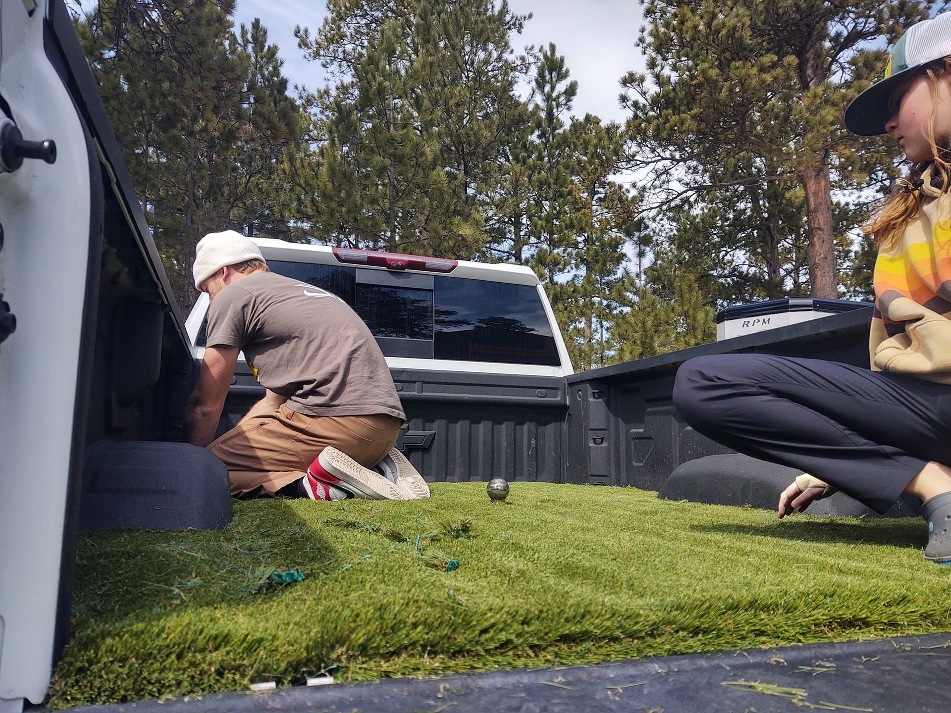 Two people work on a patch of artificial turf installed in the bed of a white pickup truck parked outdoors.